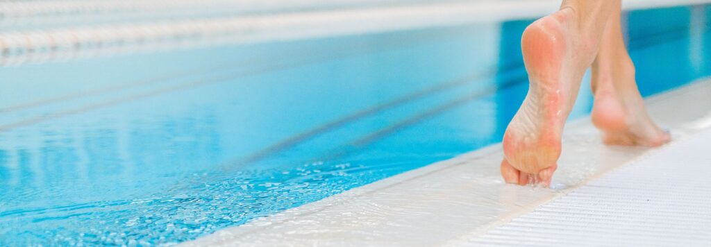 Home Close-up of bare feet walking on the wet edge of a swimming pool with clear blue water.